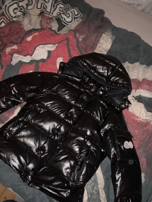 Svart glansig dunjacka från Moncler - Säljer en svart, glansig dunjacka från Moncler med huva och klassisk logga på ärmen. Jackan har en puffig design, flera fickor och är tillverkad i ett blankt material som ger en cool look. Perfekt för kalla vinterdagar och riktigt snygg till streetwear. Det är en skada på jackan men inte så farlig, det enda med skadan är att det kan se lite konstigt ut men det är inget man märker om man bara är ute. Det är lite mindre fluff pga att det har flugit ut lite innan vi sydde. Men det är bara o skriva.