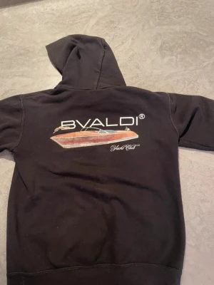 Svart BVALDI hoodie med båttryck - Svart hoodie från BVALDI med stor färgglad båtgrafik och logga på ryggen samt mindre logga och texten 'Yacht Club' på bröstet. Klassisk huva och känguruficka framtill. Perfekt för dig som gillar marina vibbar och streetwear.