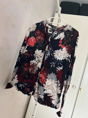 Blommig långärmad blus i mörka toner - Snygg blus med blommigt mönster i vinrött, vitt och svart. Blusen har långa ärmar, v halsringning med liten knappdetalj framtill och är tillverkad i ett tunt, luftigt material. Perfekt för dig som gillar en romantisk och trendig stil.