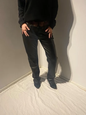 Svarta raka byxor i skinnimitation - Säljer ett par svarta byxor i skinnimitation med rak passform och hög midja. Byxorna har klassiska fickor både fram och bak samt bälteshällor. Snygga att styla med en enkel tröja eller kavaj för en trendig look.