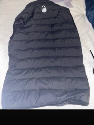 Svart dunväst från Sail Racing - Snygg svart dunväst från Sail Racing med quiltad design och dragkedja framtill. Västen har hög krage och två sidofickor med dragkedja. Perfekt för lager-på-lager och streetwear-stil. Diskret logga på bröstet och ryggen.