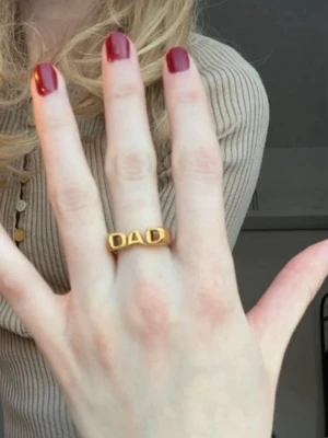 Guldfärgad ring i rostfritt stål med DAD-text - Cool ring i guldfärgad metall med texten 'DAD' i stora bokstäver. Ringen har en chunky och modern design som sticker ut och passar perfekt för dig som vill ha ett statement-smycke. Perfekt accessoar för att visa din stil. Den är i rostfritt stål och tål vatten.