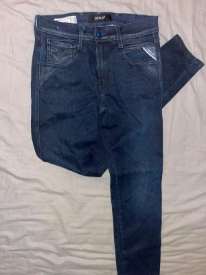 Mörkblå Replay Hyperflex jeans - Snygga mörkblå jeans från Replay Hyperflex med klassisk femficksdesign och stretchigt material. Jeansen har smal passform, blå detaljer vid fickorna och läderpatch med logga bak i midjan. Perfekta för en trendig och bekväm look. Säljs för att jag inte använder dem.