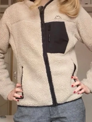 Beige teddyjacka från Kappa - Mysig beige teddyjacka från Kappa med svart bröstficka och dragkedja framtill. Jackan har svarta detaljer och en broderad Kappa-logga på bröstet. Perfekt för kalla dagar när du vill vara både varm och snygg.