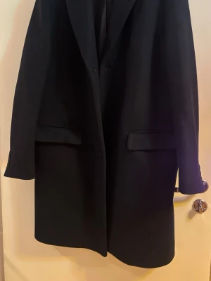 Svart ullkappa från COS - Stilren svart kappa från COS i klassisk rak modell. Kappan har två fickor framtill med lock och knäppning framtill. Perfekt för dig som gillar minimalistisk design och vill ha en tidlös look. Materialet är ullblandning som håller dig varm under kalla dagar.