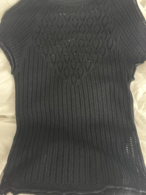 Svart stickad topp med hålmönster - Snygg svart stickad topp med korta ärmar och detaljerat hålmönster framtill. Toppen har en figurnära passform och är tillverkad i ett tunt, luftigt material som ger en cool look. Perfekt att styla lager på lager eller som den är.