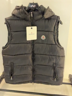Svart dunväst från Moncler - Stilren svart dunväst från Moncler med klassisk logotyp på bröstet. Västen har två dragkedjefickor framtill, hög krage och avtagbar huva. Tillverkad i glansigt material med quiltad design och dragkedja hela vägen upp.