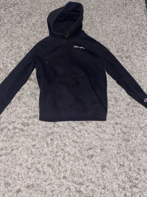 Svart Champion hoodie med logga - Svart hoodie från Champion med klassisk logga på bröstet och ärmen. Hoodien har en stor känguruficka framtill och ribbade muddar vid ärmslut och nederkant. Perfekt för en avslappnad och sportig stil.