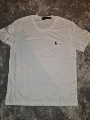 Vit t-shirt från Polo Ralph Lauren - Klassisk vit t-shirt från Polo Ralph Lauren med den ikoniska svarta polospelaren broderad på bröstet. T-shirten har rund hals och är tillverkad i mjuk bomull som känns skön mot huden. Perfekt basplagg för en clean och stilren look. Storlek M