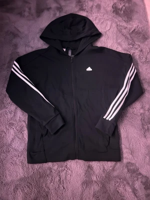 Svart hoodie med dragkedja från Adidas - Säljer en svart hoodie från Adidas med dragkedja och klassiska vita ränder längs ärmarna. Tröjan har huva och ribbade muddar vid ärmslut och nederkant. Adidas-logga på bröstet. Perfekt för en sportig och avslappnad stil.