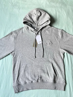 Grå hoodie från Dior med logga - Snygg grå hoodie från Dior med broderad CD-logga på bröstet. Klassisk modell med huva och dragsnören samt en stor magficka framtill. Tillverkad i mjukt bomullsmaterial som känns skönt mot huden. Perfekt för en avslappnad och trendig look.