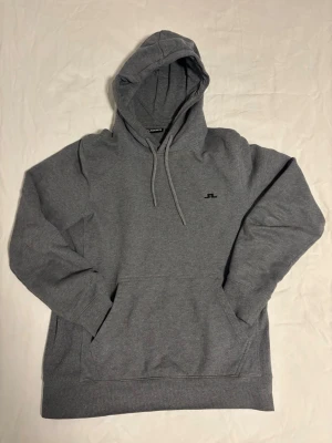 J.Lindeberg Hoodie - Klassisk grå hoodie från J.Lindeberg med liten svart logga på bröstet. Hoodien har justerbar huva med snörning, magficka och ribbade muddar vid ärmar och nederkant. Tillverkad i mjuk bomull för skön känsla.
