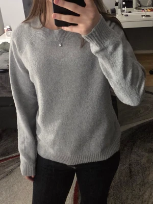 Grå stickad tröja från VERA MODA - Mysig grå stickad tröja från VERA MODA med rund halsringning och långa ärmar. Tröjan har ribbstickade muddar vid ärmslut och nederkant. Perfekt för lager-på-lager och enkel att matcha med olika outfits.