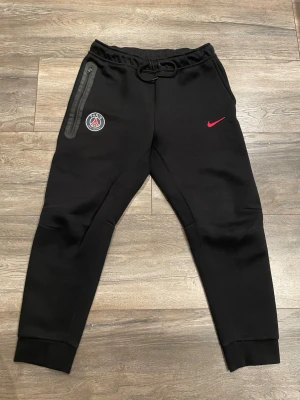 Nike tech PSG - Säljer ett par svarta mjukisbyxor från Nike i samarbete med PSG. Byxorna har en röd Nike-logga och PSG-märke på vänster ben, dragkedjeförsedd ficka och resår i midjan med snörning. Tillverkade i mjukt material och har ribbade muddar vid bensluten.