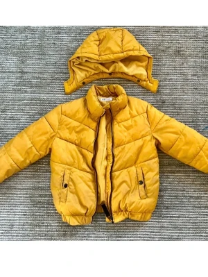 Gul pufferjacka från H&M med huva - Gul pufferjacka från H&M i storlek 140 med avtagbar huva och två fickor med knappstängning. Jackan har dragkedja framtill och elastiska muddar vid ärmslut och nederkant. Har fått en färgfläck vid ena mudden, se bild. 