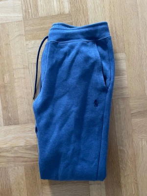 Blå mjukisbyxor från Polo Ralph Lauren - Blå mjukisbyxor från Polo Ralph Lauren med dragsko i midjan och diskret logga broderad på benet. Tillverkade i mjukt bomullsmaterial för maximal komfort. Klassisk passform med fickor på sidorna och elastisk midja.