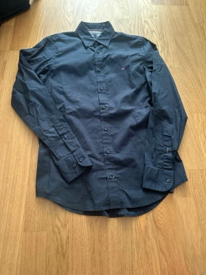 Mörkblå skjorta Tommy Hilfiger S - Snygg mörkblå skjorta från Tommy Hilfiger i slim fit med klassisk krage och små broderade loggan på bröstet. Skjortan har långa ärmar och knappar hela vägen framtill. Tillverkad i mjuk bomull med lite stretch för extra komfort.
