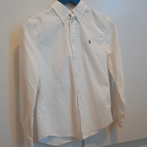 Vit skjorta från Ralph Lauren - Klassisk vit skjorta från Ralph Lauren med broderad logga på bröstet. Skjortan har lång ärm, knappar framtill och en snygg krage. Custom fit-modell i mjuk bomull som passar perfekt till jeans eller chinos.
