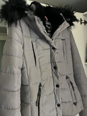 Grå pufferjacka med svart fuskpäls - Säljer en grå pufferjacka från Marikoo med svart fuskpäls på huvan. Jackan har dragkedjor och knappar framtill samt flera fickor. Insidan är fodrad med mjuk svart fleece. Perfekt för kalla dagar. 