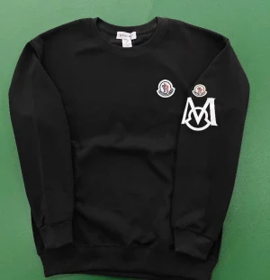 Svart sweatshirt från Moncler - Svart sweatshirt från Moncler med rund halsringning och ribbade muddar. Tröjan har Moncler-logga på bröstet och ärmen samt en stor vit M-brodyr på vänster ärm. Perfekt för dig som gillar stilrena och exklusiva plagg.