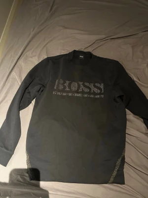 Svart BOSS sweatshirt med logga - Säljer en svart sweatshirt från BOSS med stor logga framtill i diskreta prickar. Tröjan har rund halsringning, långa ärmar och ribbade muddar. Snygga detaljer längs sidorna i samma prickiga stil som loggan. Perfekt för dig som gillar stilrena plagg med en twist.