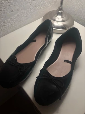 Svarta ballerinaskor med rosett från Next - Säljer ett par klassiska svarta ballerinaskor från Next i storlek 40. Skorna har en söt rosettdetalj framtill och rund tå. Yttermaterialet är glansigt syntetmaterial och innersulan är ljusrosa. Perfekta till många olika outfits!