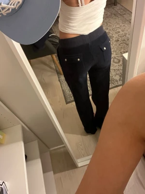 Juicy Couture byxor  - Säljer ett par mörkblå mjukisbyxor med bred midja och snörning framtill. Byxorna har raka ben, två fickor med lock och silverfärgade knappar bak samt stora fickor fram. Materialet är mjukt och känns som sammet, perfekt för chill dagar.