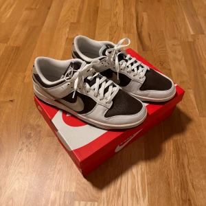 Nike Dunk Low brun och vit - Nike Dunk Low sneakers i brun och vit med klassisk snörning och rund tå. Ovandelen är i skinn med perforerade detaljer på tån och vit sula. Snygg kontrast mellan de mörka och ljusa partierna, perfekt för dig som gillar stilrena sneakers. Använda endast en gång. Annars helt nya med box. Köpta på Nike i Sthlm 