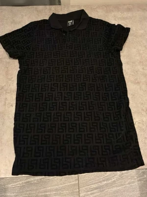 Svart mönstrad piké från Black Socks - Säljer en svart pikétröja från Black Socks i storlek XS. Tröjan har ett diskret geometriskt mönster över hela tyget, klassisk krage och knappar framtill. Perfekt för dig som gillar stilrena detaljer och vill ha något unikt i garderoben.