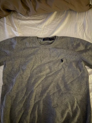 Grå stickad tröja från Polo Ralph Lauren - Klassisk grå stickad tröja från Polo Ralph Lauren i storlek M. Tröjan har rund halsringning, ribbade muddar och den ikoniska broderade loggan på bröstet. Perfekt för lager på lager under höst och vinter.