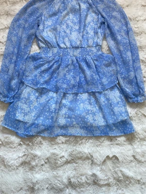 Blommig blå kortklänning Kappahl - Supersöt blå kortklänning från Kappahl med vitt blommönster. Klänningen har långärmade ärmar, smock i midjan och volangdetaljer. Tillverkad i ett lätt och luftigt material med innerfoder för extra komfort. Perfekt för sommarens alla tillfällen! Pris 40kr 