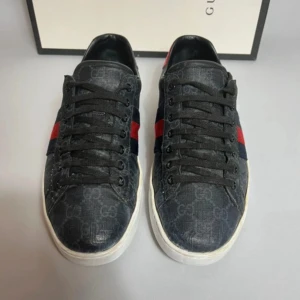 Gucci ace i svart - Snygga svarta Gucci sneakers med klassiskt GG-monogram över hela skon och ikoniska röd/blå ränder på sidorna. Skorna har svarta snören, vit platt sula och är tillverkade i canvas med skinninslag. Perfekta för dig som vill sticka ut med lyxig streetstyle.