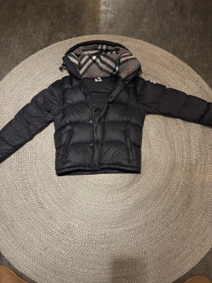 Svart pufferjacka från Burberry - Säljer en svart pufferjacka från Burberry i storlek M. Jackan har klassiskt rutigt foder i huvan, dragkedja och knappar framtill samt två fickor med dragkedja. Perfekt för kalla dagar och riktigt snygg till alla outfits.