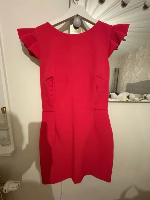 Röd kort klänning från Zara Basic - Snygg röd kort klänning från Zara Basic med volangärmar och stor rosett i ryggen. Klänningen har öppen rygg, dragkedja bak och är tillverkad i ett mjukt polyestertyg. Längden är ca 84 cm. Perfekt för dig som vill sticka ut med en färgstark look.