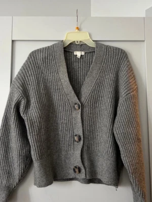 Grå ribbad kofta med knappar - Säljer en grå ribbstickad kofta med stora knappar framtill. Koftan har v-ringad hals och långa ärmar med lite loose passform. Perfekt att slänga på sig när det är kyligt ute. Materialet känns mjukt och stickat, vilket gör den extra skön.