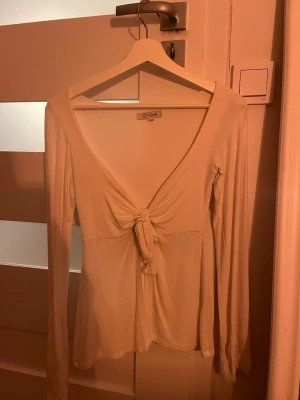 Vit långärmad topp med knyt fram - Säljer en vit långärmad topp från Design by si med djup v-ringning och snygg knytning framtill. Toppen är i mjukt, stretchigt material och har en figurnära passform som framhäver siluetten. Perfekt att styla med jeans eller kjol.