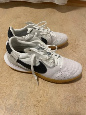 Nike streetgato futsalskor - Säljer ett par vita och svarta Nike streetgato med klassisk svart swoosh på sidan. Skorna har snörning, rund tå och platt sula. Ovandelen är i mesh och mocka med perforerade detaljer för extra ventilation. Yttersulan är i gummi med bra grepp för inomhusspel. Skorna är använda 1 gäng.