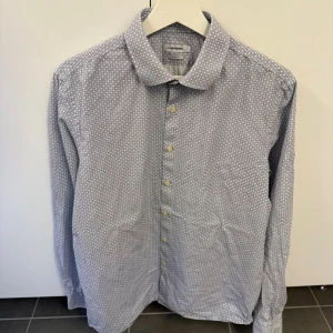 Mönstrad skjorta från J.Lindeberg - Snygg skjorta från J.Lindeberg i vitt och blått med ett diskret cirkelmönster. Skjortan har klassisk krage, långa ärmar och knäppning framtill. Tillverkad i mjuk bomull och sitter tajt med slim fit-passform. Storlek L