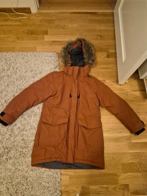 Brun parkas från Didriksons med fuskpäls - Säljer en brun parkas från Didriksons med huva och fuskpäls. Jackan är medel lång, har stora fickor framtill, två fickor vid uppe vid bröstet och är fodrad med grått innerfoder och beige teddyfoder upptill. 100% vattentät och perfekt för kalla dagar alltså den är en jättebra vinterjacka. Stängs med dragkedja och kardborre vid ärmslut. Jag har köpt den för 800 kr. Den säljs för priset den är satt på.