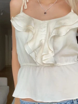 Krämvit linnetopp med volang - Säljer en krämvit linnetopp med bred volang framtill och tunna axelband som knyts på axlarna. Toppen har en lätt glansig yta och smock i midjan för en feminin siluett. Perfekt till jeans eller kjol för en fräsch sommarlook.
