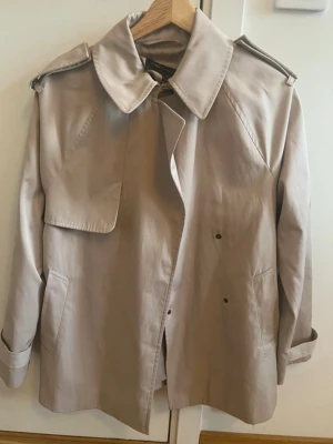 Beige trenchcoat från M&S Collection - Snygg beige trenchcoat från M&S Collection med klassisk krage, axelklaffar och dubbelknäppning. Jackan har två stora fickor framtill och är tillverkad i ett slätt material som känns lätt och stilrent. Kan användas som storlek M också. Är öppna med att diskutera priset!