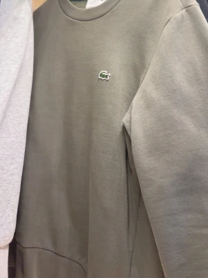 Olivgrön sweatshirt från Lacoste - Säljer en olivgrön sweatshirt från Lacoste i klassisk passform. Tröjan har rund halsringning, lång ärm och den ikoniska gröna krokodilloggan på bröstet. Tillverkad i mjuk bomull för skön vardagsstil.