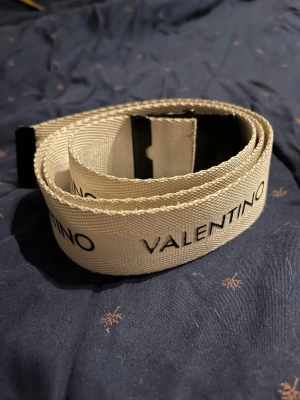 Valentino beige bälte med svart spänne - Snyggt beige bälte från Valentino med svart metallspänne och svart logotyp längs hela bandet. Bältet är gjort i slitstark syntet och har en clean, modern look som passar till många olika outfits. 100 cm i storlek 