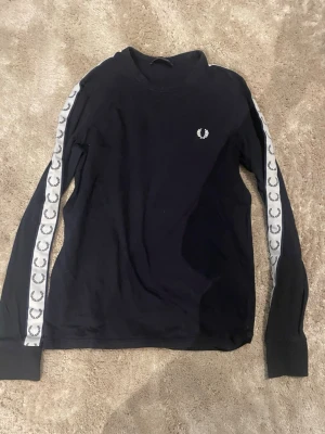 Svart långärmad tröja Fred Perry - Svart långärmad tröja från Fred Perry med vita loggor längs ärmarna och broderad logga på bröstet. Klassisk crewneck och ribbade muddar. Perfekt till streetwear-stilen.