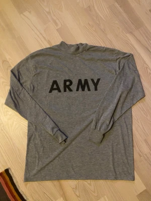 Grå långärmad ARMY t-shirt - Säljer en grå långärmad t-shirt med svart ARMY-tryck på framsidan och en stor svart grafisk detalj på ryggen. Tröjan har en enkel, rak passform och är gjord i mjukt bomullsmaterial. Perfekt för dig som gillar minimalistisk och street-inspirerad stil.