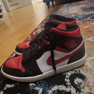 Nike Air Jordan 1 svart/röd/vit - Säljer ett par Nike Air Jordan 1 sneakers i klassisk svart, röd och vit färg. Skorna har högt skaft, svart snörning och den ikoniska Air Jordan-loggan på sidan. Ovandelen är i skinn och de har en vit sula med röd swoosh. Perfekta för dig som gillar streetwear och sneakers.