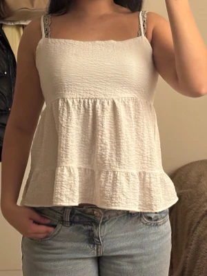 Volang toop - Supersöt vit linnetopp i storlek 38. Toppen har smala axelband, volang nertill och en snygg snörning i ryggen. Materialet är lätt och lite skrynkligt, perfekt för varma dagar. Passar till jeans eller kjol för en chill sommarlook.