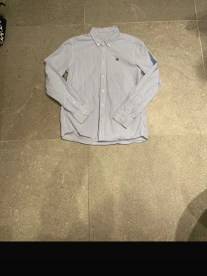 Blårandig skjorta United Colors of Benetton 152 cm - Säljer en klassisk blårandig skjorta från United Colors of Benetton. Skjortan har vit knappstängning, lång ärm och en snygg broderad logga på bröstet. Perfekt till jeans eller chinos för en clean och fräsch look.