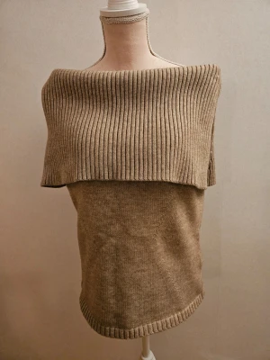 Beige ribbad stickad tröja från Adoore - Säljer en beige stickad tröja från Adoor med bred ribbad krage som ger en snygg offshoulder-look. Tröjan är i mjukt material och har ribbade detaljer vid nederkant och hals. Perfekt för lager på lager under höst och vinter.