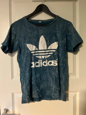 Blå Adidas t-shirt med stor logga - Säljer en blå t-shirt från Adidas med stor vit Trefoil-logga på bröstet. T-shirten har korta ärmar och en tvättad, lite vintage look. Perfekt för dig som gillar streetwear och vill ha något avslappnat men ändå snyggt.